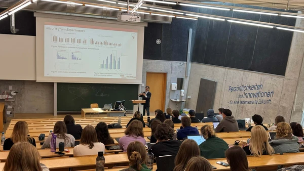 View from the back of the lecture hall, a presentation on the screen at the front, a speaker in a suit at the lectern, the students can be seen from behind  